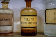 Three vintage glass bottles with old labels are displayed. The central bottle has a yellowish tint and is labeled 'SOLUTIO MERCUROCHROM. 20%'. The bottles to the left and right are partially visible, with one labeled 'EUCALYPTI'. The design and typography on the labels give a historic and medicinal feel.