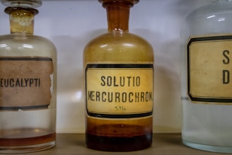 Three vintage glass bottles with old labels are displayed. The central bottle has a yellowish tint and is labeled 'SOLUTIO MERCUROCHROM. 20%'. The bottles to the left and right are partially visible, with one labeled 'EUCALYPTI'. The design and typography on the labels give a historic and medicinal feel.
