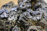 Cluster of shiny, black crystals with geometric shapes and defined edges, intertwined with some earthy, brown substances. The crystals have a glossy, reflective surface.