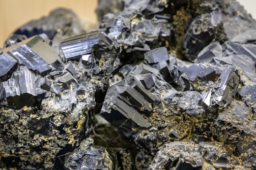 Cluster of shiny, black crystals with geometric shapes and defined edges, intertwined with some earthy, brown substances. The crystals have a glossy, reflective surface.