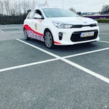 A compact white car parked in a mostly empty parking lot. The vehicle features red and blue patterned designs on its side, a roof sign indicating it is a driving school car, and a visible European license plate. The background includes leafless trees and a cloudy sky.