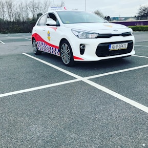 A compact white car parked in a mostly empty parking lot. The vehicle features red and blue patterned designs on its side, a roof sign indicating it is a driving school car, and a visible European license plate. The background includes leafless trees and a cloudy sky.