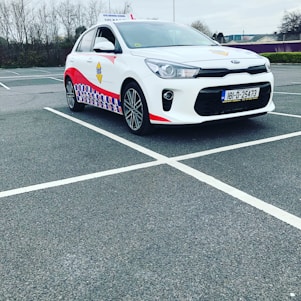 A compact white car parked in a mostly empty parking lot. The vehicle features red and blue patterned designs on its side, a roof sign indicating it is a driving school car, and a visible European license plate. The background includes leafless trees and a cloudy sky.