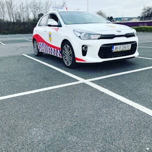 A compact white car parked in a mostly empty parking lot. The vehicle features red and blue patterned designs on its side, a roof sign indicating it is a driving school car, and a visible European license plate. The background includes leafless trees and a cloudy sky.