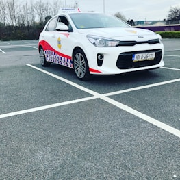 A compact white car parked in a mostly empty parking lot. The vehicle features red and blue patterned designs on its side, a roof sign indicating it is a driving school car, and a visible European license plate. The background includes leafless trees and a cloudy sky.