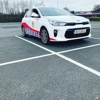 A compact white car parked in a mostly empty parking lot. The vehicle features red and blue patterned designs on its side, a roof sign indicating it is a driving school car, and a visible European license plate. The background includes leafless trees and a cloudy sky.