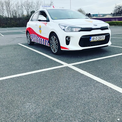 A compact white car parked in a mostly empty parking lot. The vehicle features red and blue patterned designs on its side, a roof sign indicating it is a driving school car, and a visible European license plate. The background includes leafless trees and a cloudy sky.