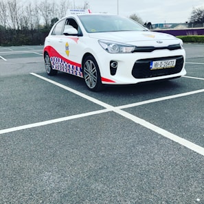 A compact white car parked in a mostly empty parking lot. The vehicle features red and blue patterned designs on its side, a roof sign indicating it is a driving school car, and a visible European license plate. The background includes leafless trees and a cloudy sky.