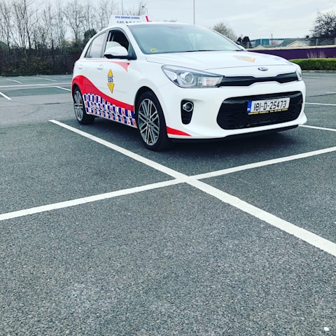 A compact white car parked in a mostly empty parking lot. The vehicle features red and blue patterned designs on its side, a roof sign indicating it is a driving school car, and a visible European license plate. The background includes leafless trees and a cloudy sky.