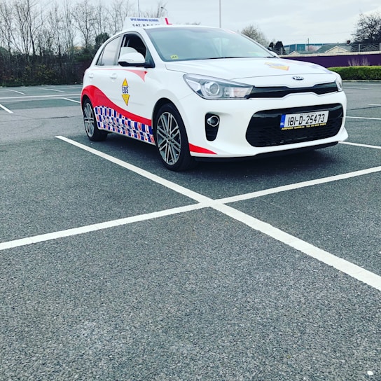 A compact white car parked in a mostly empty parking lot. The vehicle features red and blue patterned designs on its side, a roof sign indicating it is a driving school car, and a visible European license plate. The background includes leafless trees and a cloudy sky.