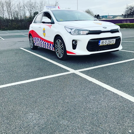A compact white car parked in a mostly empty parking lot. The vehicle features red and blue patterned designs on its side, a roof sign indicating it is a driving school car, and a visible European license plate. The background includes leafless trees and a cloudy sky.