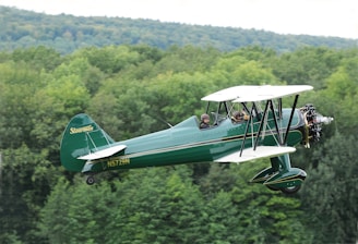 A vintage biplane with a distinct green body and white accents is flying over a lush forested area. Two individuals are seated in the open cockpit, wearing aviator goggles and helmets. The aircraft's propeller is visibly in motion, and the background features a range of green trees under a cloudy sky.