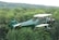 A vintage biplane with a distinct green body and white accents is flying over a lush forested area. Two individuals are seated in the open cockpit, wearing aviator goggles and helmets. The aircraft's propeller is visibly in motion, and the background features a range of green trees under a cloudy sky.