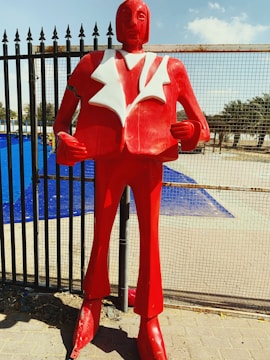 A large red statue of a person wearing a suit with exaggerated shoulders and a stylized design. The suit has a white lapel, and the figure stands upright with its hands positioned as if holding onto its jacket. The background features a metal fence and a blue tarp, with some palm trees visible in the distance under a clear sky.