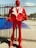 A large red statue of a person wearing a suit with exaggerated shoulders and a stylized design. The suit has a white lapel, and the figure stands upright with its hands positioned as if holding onto its jacket. The background features a metal fence and a blue tarp, with some palm trees visible in the distance under a clear sky.