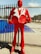 A large red statue of a person wearing a suit with exaggerated shoulders and a stylized design. The suit has a white lapel, and the figure stands upright with its hands positioned as if holding onto its jacket. The background features a metal fence and a blue tarp, with some palm trees visible in the distance under a clear sky.
