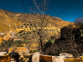 A mountainous landscape with arid slopes covered in patches of vegetation. Several houses with yellow and earthy tones are nestled within the hillside. In the foreground, brightly colored textiles are draped across a railing. The sky above is clear and deep blue.