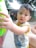 A young child with short hair wearing a sleeveless shirt reaching out towards a large green object. The background shows an outdoor setting with other people in a shaded area under a roof made of bamboo or similar material. The scene appears casual and relaxed.