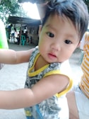 A young child with short dark hair, wearing a sleeveless top with yellow trim, is close to the camera. In the background, there are several people sitting around a table in an outdoor setting with a rustic shelter.