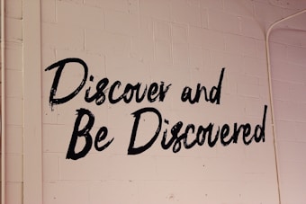 Black text reading 'Discover and Be Discovered' is painted on a white, textured brick wall. The writing style is bold and casual.