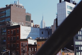 An urban skyline featuring several buildings of varying heights and architectural styles. A prominent structure with Arabic and English text is visible, stating the ownership of a book by Fathallah Saad from March 1892. In the background, a famous skyscraper rises above the other buildings. The scene is viewed through a diagonal metallic railing or beam.