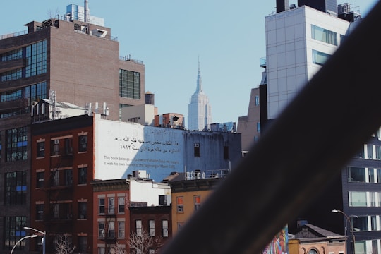 An urban skyline featuring several buildings of varying heights and architectural styles. A prominent structure with Arabic and English text is visible, stating the ownership of a book by Fathallah Saad from March 1892. In the background, a famous skyscraper rises above the other buildings. The scene is viewed through a diagonal metallic railing or beam.