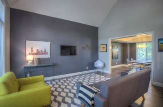 A modern living room with a high ceiling, featuring a gray accent wall adorned with a colorful cityscape artwork and a wall-mounted TV. The room includes a vibrant lime green armchair and a gray sofa positioned on a geometric patterned rug. A small console table with a unique lamp sits to the side. Through an open doorway, a dining area with a rectangular table and chairs can be seen, enhanced by natural light from large windows.