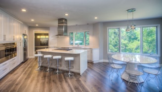 A modern kitchen features sleek white cabinetry and a spacious island with four bar stools. The countertops are white and the kitchen includes stainless steel appliances, such as an oven and refrigerator. Large windows offer a view of lush greenery outside, while a round dining table with wire chairs sits nearby. Pendant lighting fixtures add a stylish touch to the ceiling. The flooring is a dark wood, complementing the overall airy and bright atmosphere.