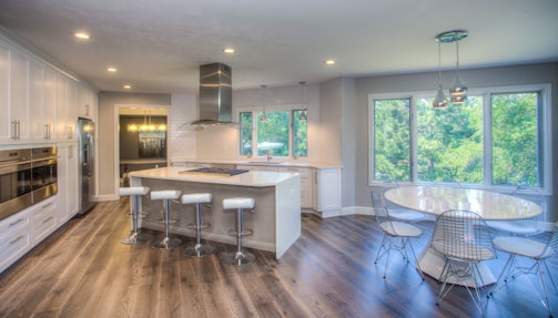 A modern kitchen features sleek white cabinetry and a spacious island with four bar stools. The countertops are white and the kitchen includes stainless steel appliances, such as an oven and refrigerator. Large windows offer a view of lush greenery outside, while a round dining table with wire chairs sits nearby. Pendant lighting fixtures add a stylish touch to the ceiling. The flooring is a dark wood, complementing the overall airy and bright atmosphere.