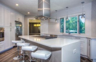 A modern kitchen features sleek white cabinetry and a central island with a gas stove and four white bar stools. Stainless steel appliances, including a fridge and oven, complement the minimalistic style. Three pendant lights hang above the island, and large windows reveal a green, tree-filled view outside. The floor is made of dark wood, adding contrast to the bright interior.