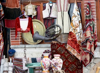 A collection of traditional textiles and clothing items, with intricately designed rugs hanging beside embroidered garments. A large brass instrument and two ornately decorated ceramic vases add to the eclectic display. The background consists of a brick wall and wooden frame, suggesting a vintage shop setting.