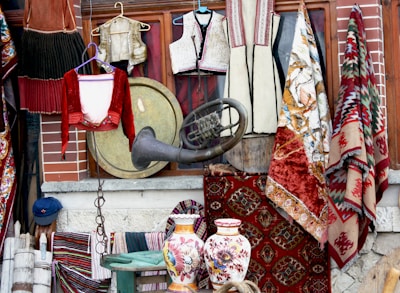 A collection of traditional textiles and clothing items, with intricately designed rugs hanging beside embroidered garments. A large brass instrument and two ornately decorated ceramic vases add to the eclectic display. The background consists of a brick wall and wooden frame, suggesting a vintage shop setting.