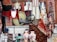 A collection of traditional textiles and clothing items, with intricately designed rugs hanging beside embroidered garments. A large brass instrument and two ornately decorated ceramic vases add to the eclectic display. The background consists of a brick wall and wooden frame, suggesting a vintage shop setting.