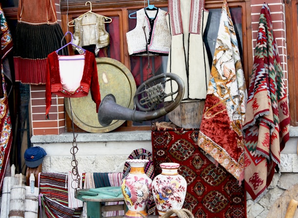 A collection of traditional textiles and clothing items, with intricately designed rugs hanging beside embroidered garments. A large brass instrument and two ornately decorated ceramic vases add to the eclectic display. The background consists of a brick wall and wooden frame, suggesting a vintage shop setting.