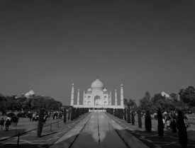 A monochromatic photograph of a large, historic marble mausoleum with a prominent dome and surrounding minarets. In the foreground, a long rectangular water feature is flanked by manicured gardens with towering cypress trees. Numerous visitors are walking along the pathways, capturing the grand architecture.