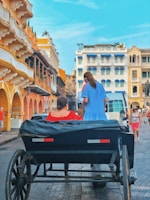 A vibrant street scene with a horse-drawn carriage carrying two passengers in colorful dresses. The buildings in the background display a mix of colonial architecture with bright facades and balconies adorned with plants. Pedestrians are visible walking along the street, and the sky is clear and blue.