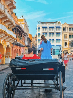 A vibrant street scene with a horse-drawn carriage carrying two passengers in colorful dresses. The buildings in the background display a mix of colonial architecture with bright facades and balconies adorned with plants. Pedestrians are visible walking along the street, and the sky is clear and blue.