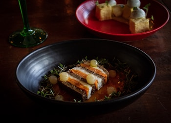 A dark ceramic bowl contains an artistic food presentation with layers of ingredients garnished with herbs and small translucent spheres. In the background, a red plate holds a dessert with cubes and domes. A green-stemmed glass is partially visible on the side, all set on a wooden table.