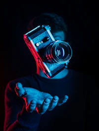 Karanlık bir ortamda kırmızı ve mavi ışıkla aydınlanmış, açık bir avucun üzerinde havada duran eski fotoğraf makinesi