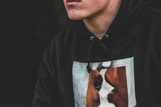 A person wearing a black hoodie with a graphic design featuring two faces on the front. The image is focused on the torso and part of the face. The hoodie has a drawstring with metal eyelets.