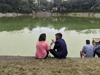 Several people are sitting by a pond with greenish water, surrounded by lush greenery. Two individuals are engaged in conversation in the foreground, one wearing a pink shirt and the other in a plaid shirt. The background features dense trees and some buildings partially visible over the treeline.