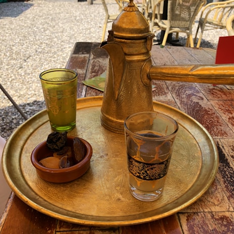 A traditional golden coffee pot is placed on a decorative brass tray. Two glasses, one with an ornate black pattern, are on the tray along with a small bowl containing dried fruits. The setting is outdoors on a wooden table, surrounded by gravel and wicker chairs.