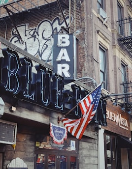 A street corner with a rustic bar and restaurant facade featuring a prominent neon sign reading 'BAR' and 'BLUE'. An American flag is displayed, accompanied by graffiti on a brick wall. The scene includes a vintage vibe with a traditional wooden storefront.