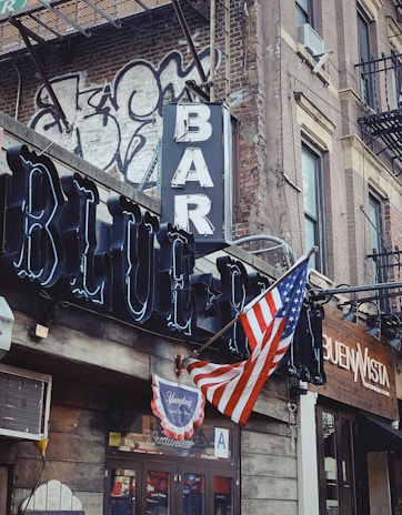 A street corner with a rustic bar and restaurant facade featuring a prominent neon sign reading 'BAR' and 'BLUE'. An American flag is displayed, accompanied by graffiti on a brick wall. The scene includes a vintage vibe with a traditional wooden storefront.