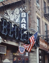 A street corner with a rustic bar and restaurant facade featuring a prominent neon sign reading 'BAR' and 'BLUE'. An American flag is displayed, accompanied by graffiti on a brick wall. The scene includes a vintage vibe with a traditional wooden storefront.