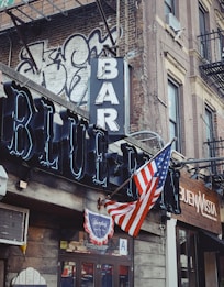 A street corner with a rustic bar and restaurant facade featuring a prominent neon sign reading 'BAR' and 'BLUE'. An American flag is displayed, accompanied by graffiti on a brick wall. The scene includes a vintage vibe with a traditional wooden storefront.
