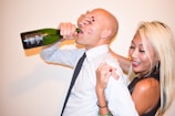 A man is drinking from a green bottle while a woman playfully covers his eyes and smiles. The man is wearing a white shirt and tie with lipstick marks on his cheek. The woman has long blonde hair and polished red nails.