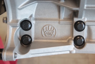 A close-up view of a metallic surface with detailed textures and patterns. Four black, round fasteners are symmetrically placed around an embossed logo featuring stylized graphics and the letters 'BM'. The metallic surface has a silver-gray color and appears industrial in nature.