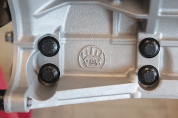 A close-up view of a metallic surface with detailed textures and patterns. Four black, round fasteners are symmetrically placed around an embossed logo featuring stylized graphics and the letters 'BM'. The metallic surface has a silver-gray color and appears industrial in nature.