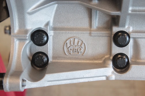 A close-up view of a metallic surface with detailed textures and patterns. Four black, round fasteners are symmetrically placed around an embossed logo featuring stylized graphics and the letters 'BM'. The metallic surface has a silver-gray color and appears industrial in nature.